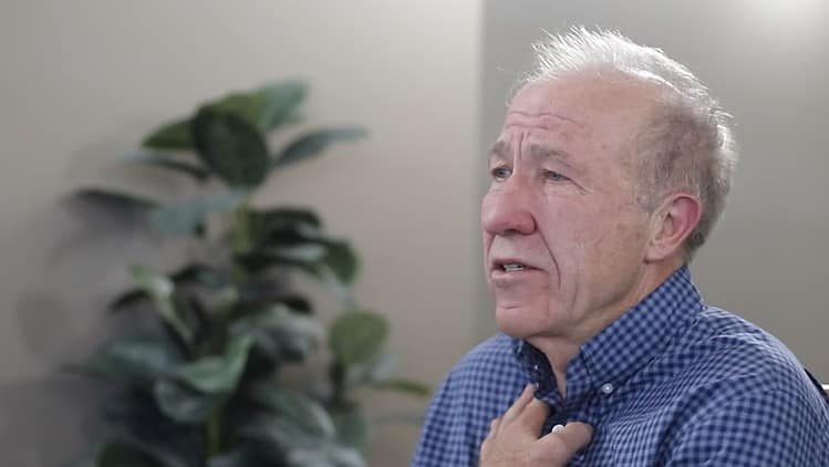 Man sitting with a hand to his chest, earnestly speaking with a plant in the background.