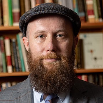 Headshot of Alan. Man in a coat & tie with a large red beard and a courier hat infront of bookcases behind him.