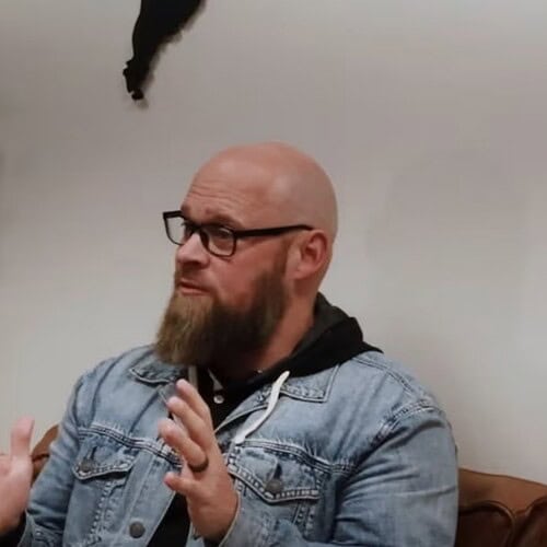 Noah Oldham seating speaking with his hands with a plant int he background