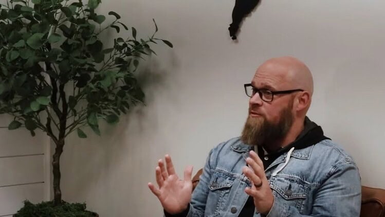Noah Oldham seating speaking with his hands with a plant int he background