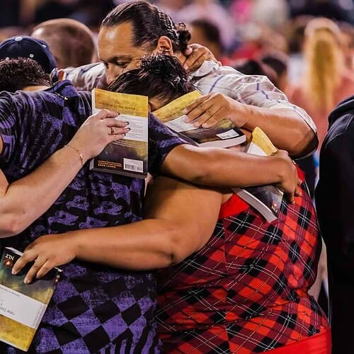 a diverser group of people with their arms around one another, with their Bibles in corporate prayer