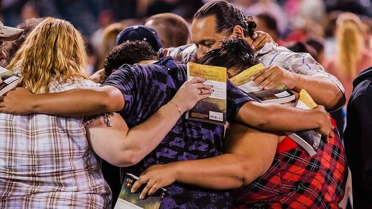 a diverser group of people with their arms around one another, with their Bibles in corporate prayer