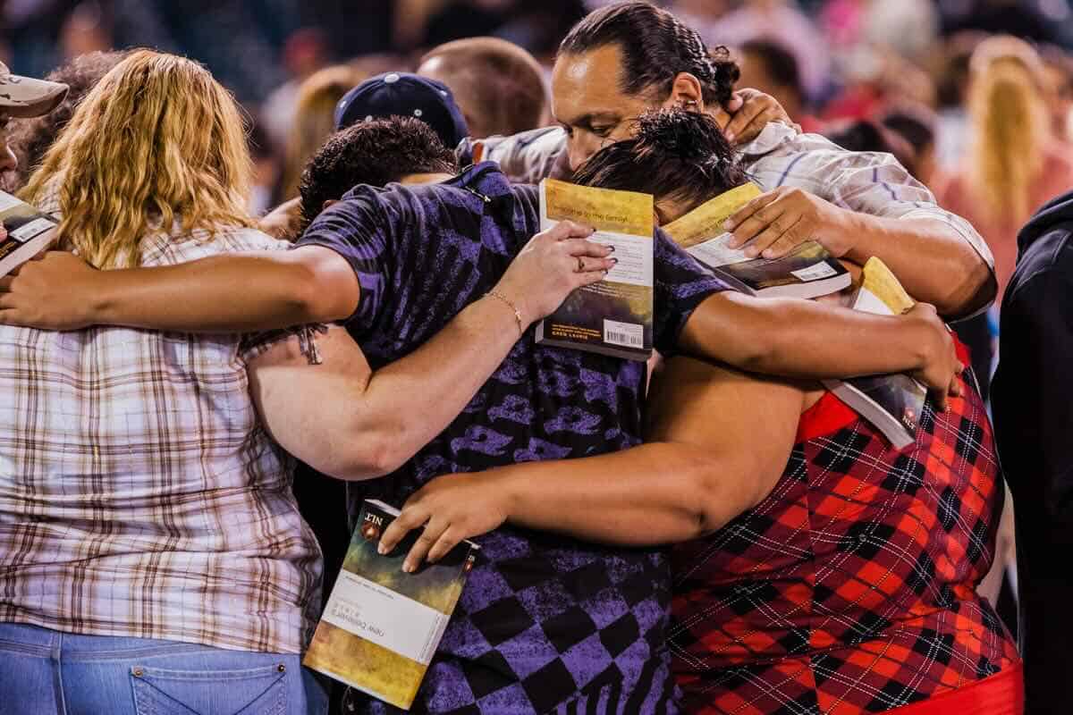 a diverser group of people with their arms around one another, with their Bibles in corporate prayer