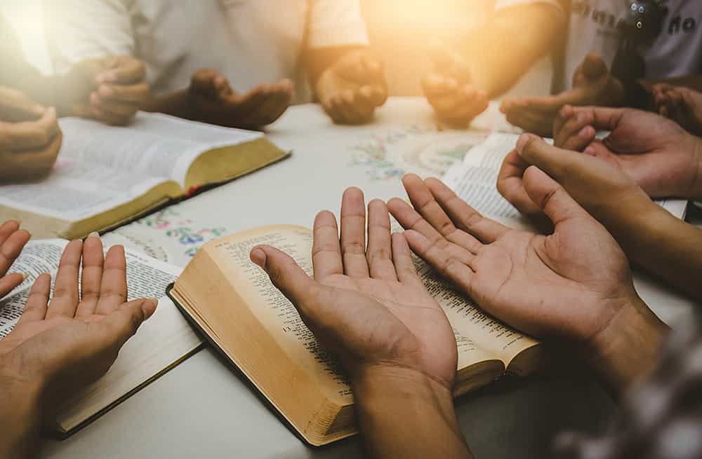 5 people around a table with hands on bibles palms up